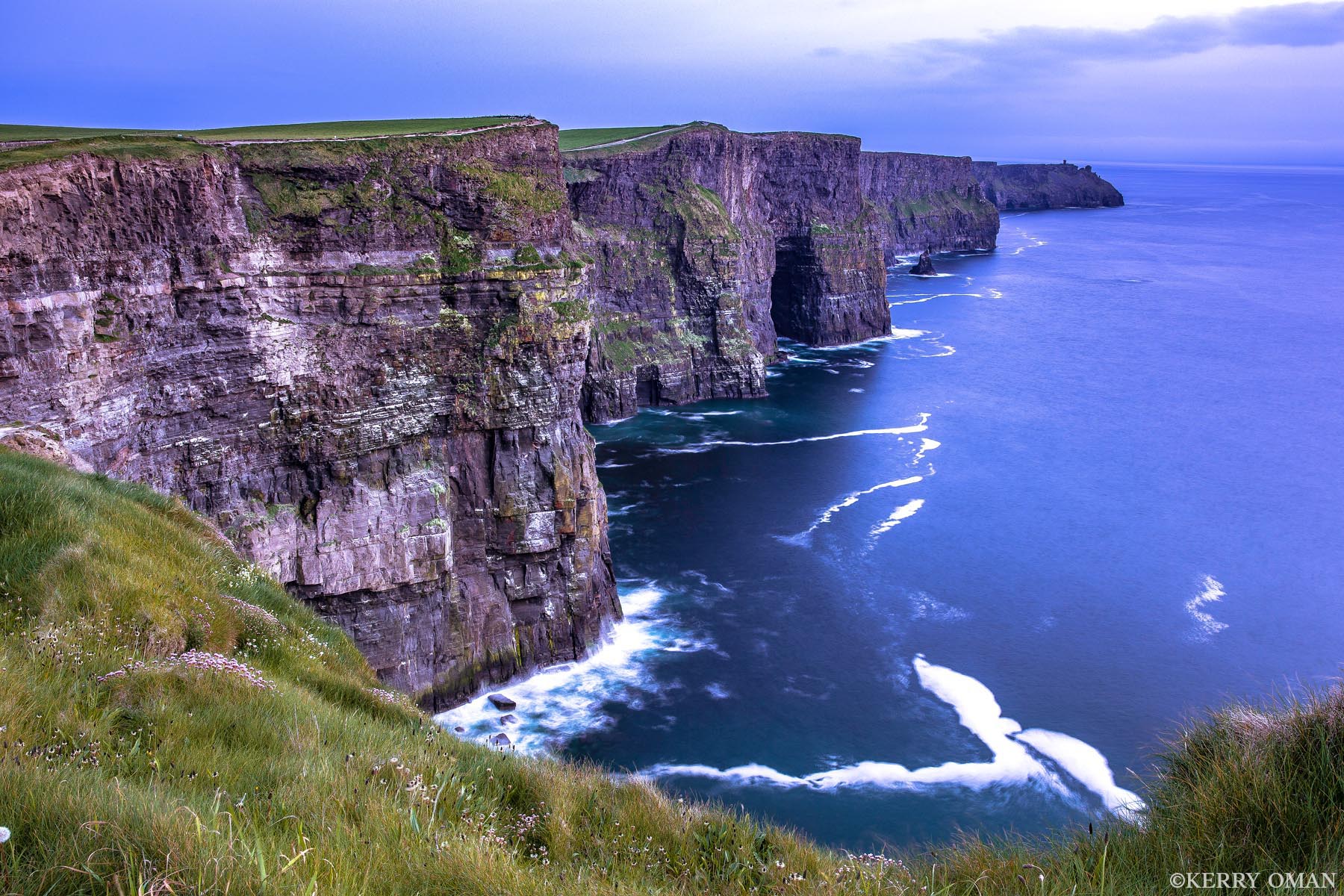 Along the Cliffs of Moher The Atlantic carves its quiet path along the base of the Cliffs of Moher, where ancient stone walls rise in rugged steps above...