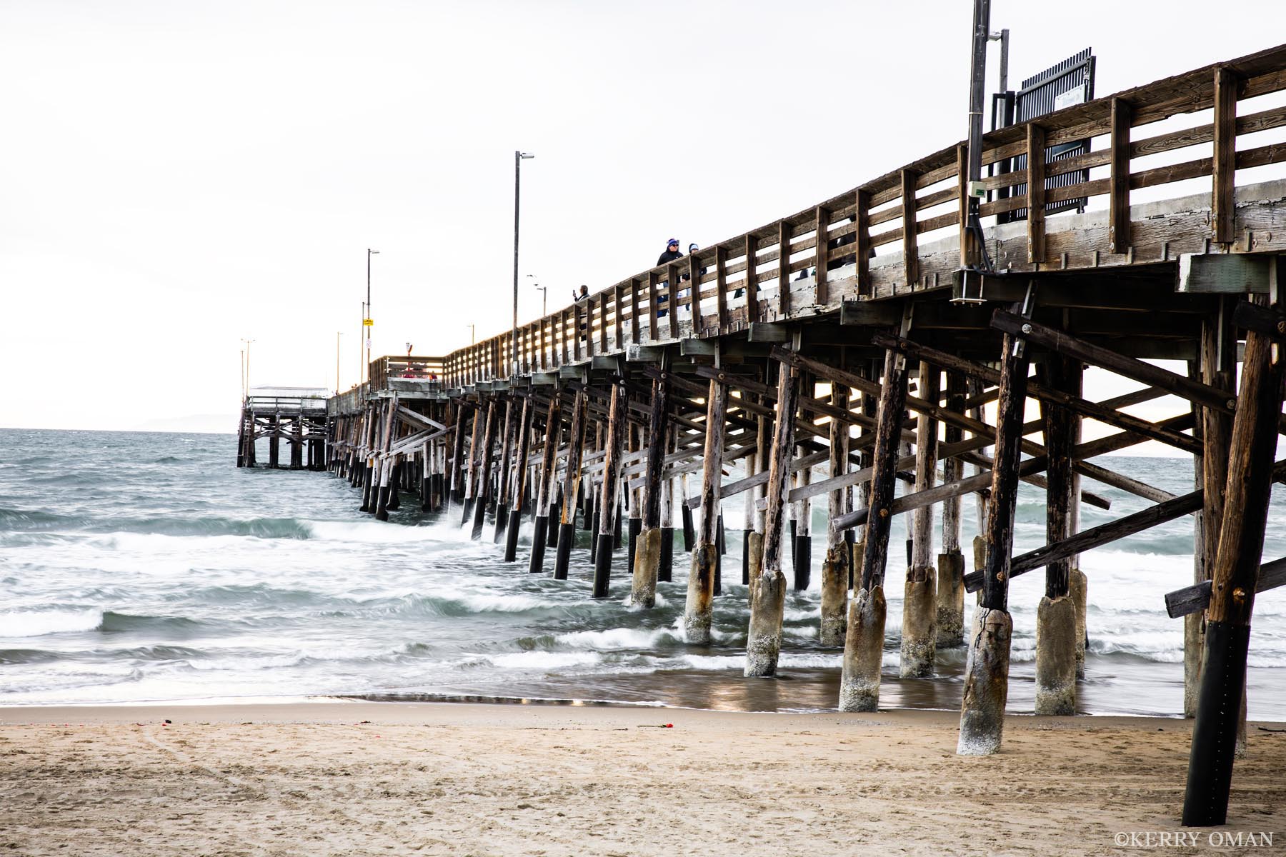 Under Newport Pier Waves roll in beneath the wooden supports of Newport Pier as beachgoers take in the view from above. Captured in late light...